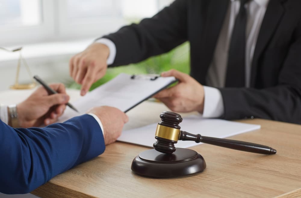 A faceless, unrecognizable male client is seated at a table, signing a paper contract while making an agreement with a lawyer. A wooden gavel is positioned prominently on the office desk, symbolizing legal authority and justice.