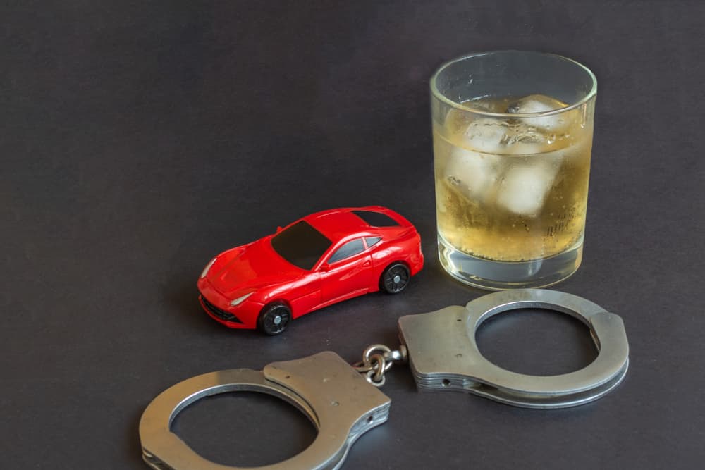 A toy car, handcuffs, and a bottle of alcohol are displayed against a colorful background, illustrating the dangers of driving under the influence.