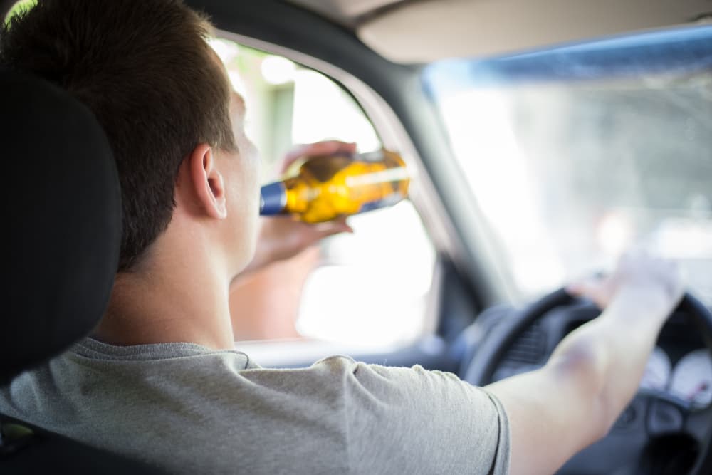 A young driver holds a bottle of alcohol while behind the wheel of a car, emphasizing the message of a campaign against drinking and driving.