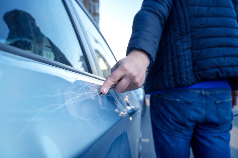 A man vandalizing a car by scratching the door with a screwdriver in a parking lot, damaging the property out of revenge for betrayal or as a form of intimidation.