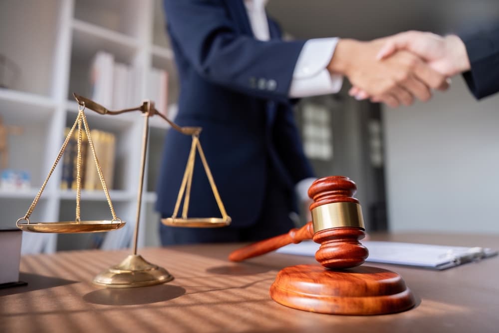 Professional handshake between lawyer and client in a business meeting, with scales of justice and a gavel on the desk, symbolizing legal agreement and partnership in a modern office setting.